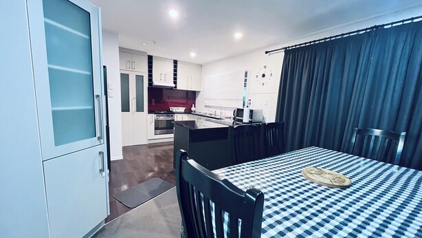 A compact kitchen with white cabinets, a microwave, and a stove. There is a dining area with a checkered table and chairs. - Holiday Home In Red Hill Papakura Auckland With Beautiful Views (Papakura)