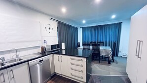 A compact kitchen with modern appliances, a dining area with a checkered tablecloth, and a view of the outdoors through curtains. - Holiday Home In Red Hill Papakura Auckland With Beautiful Views (Papakura)