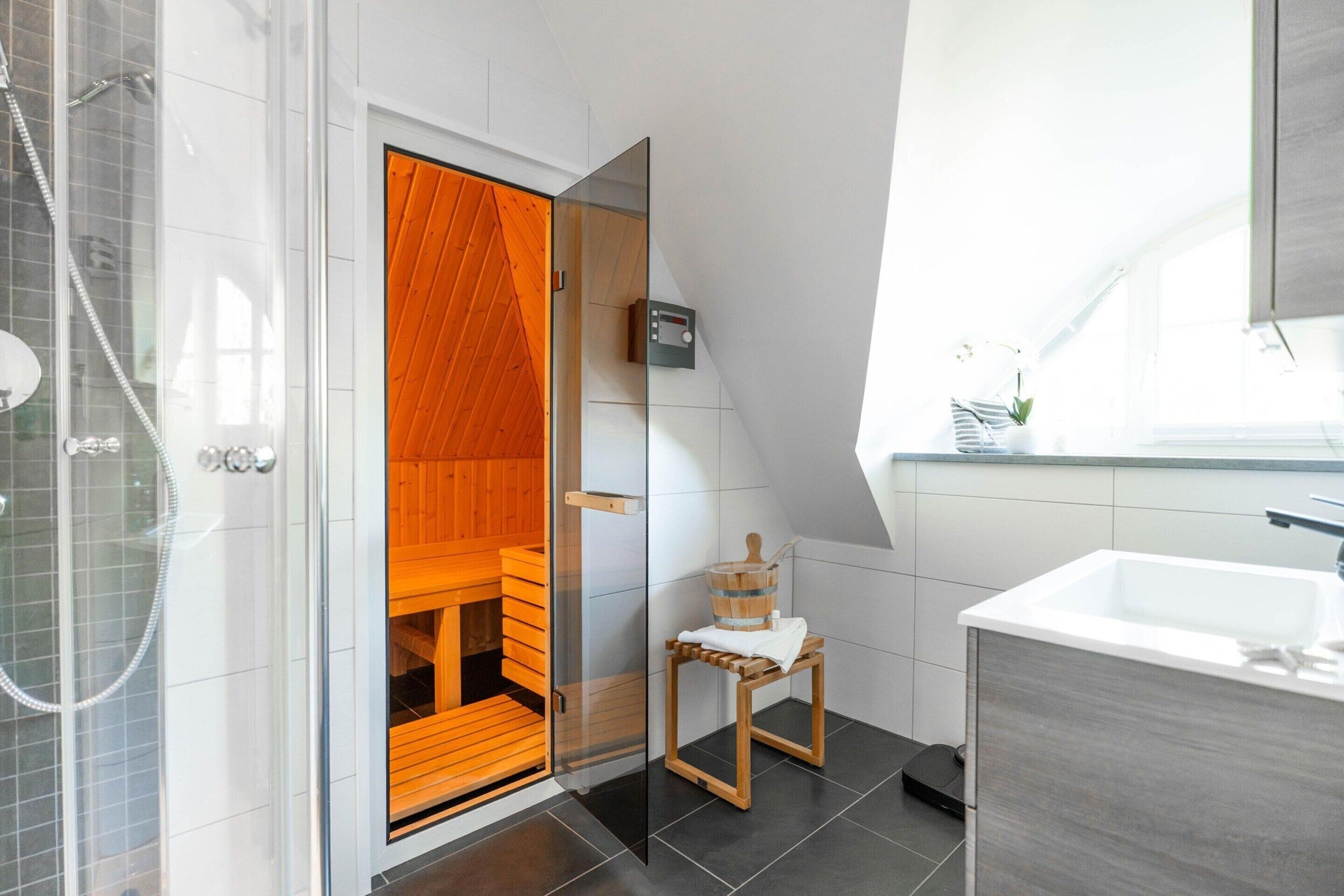 A modern bathroom with a glass shower enclosure, a wooden sauna, a small wooden stool, and a white sink.