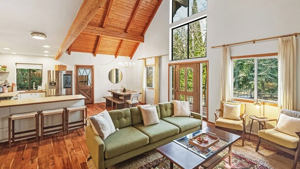 A spacious living area with a high ceiling, wooden beams, a green sofa, a dining table, and large windows offering views of greenery. - Close to Ski Slopes & Trails ~ Zoo ~ Hot Tub (Big Bear)