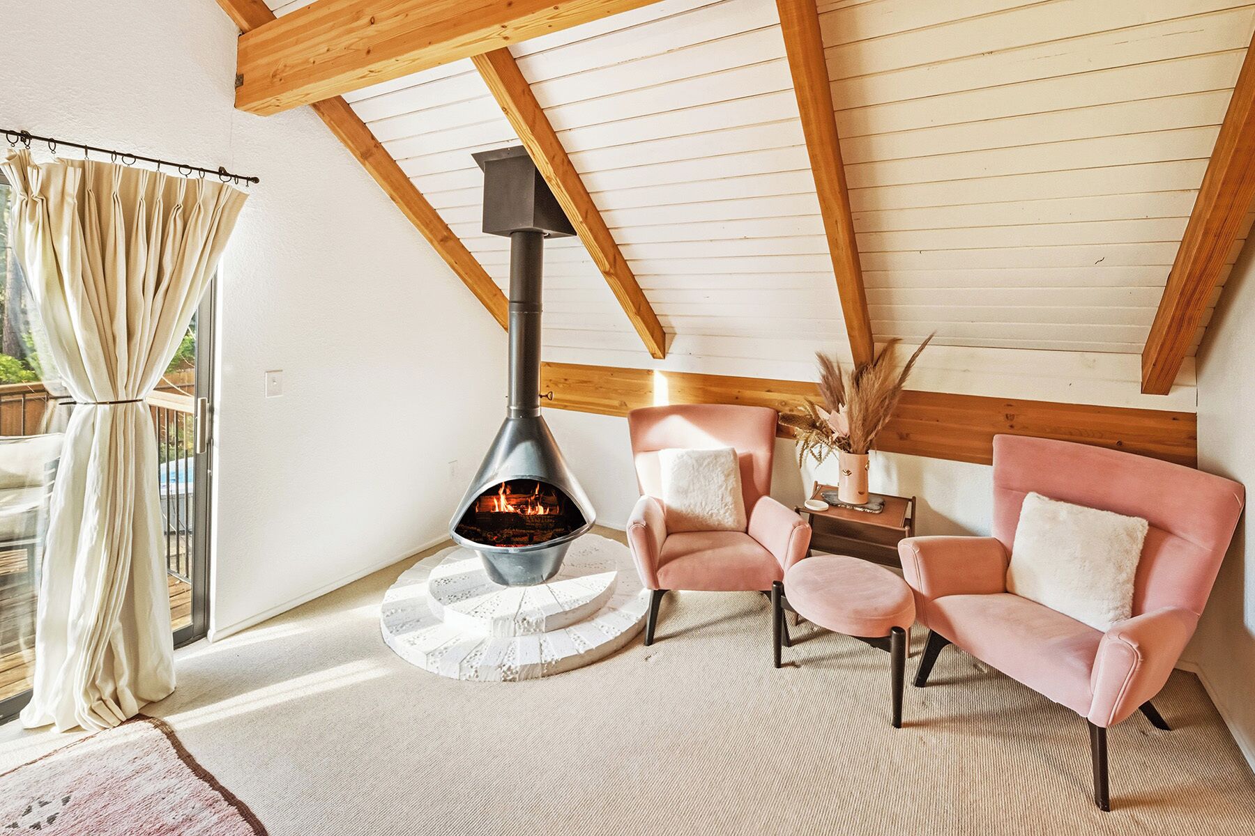 A cozy room with a wood-burning stove, two pink chairs, and a wooden ceiling.