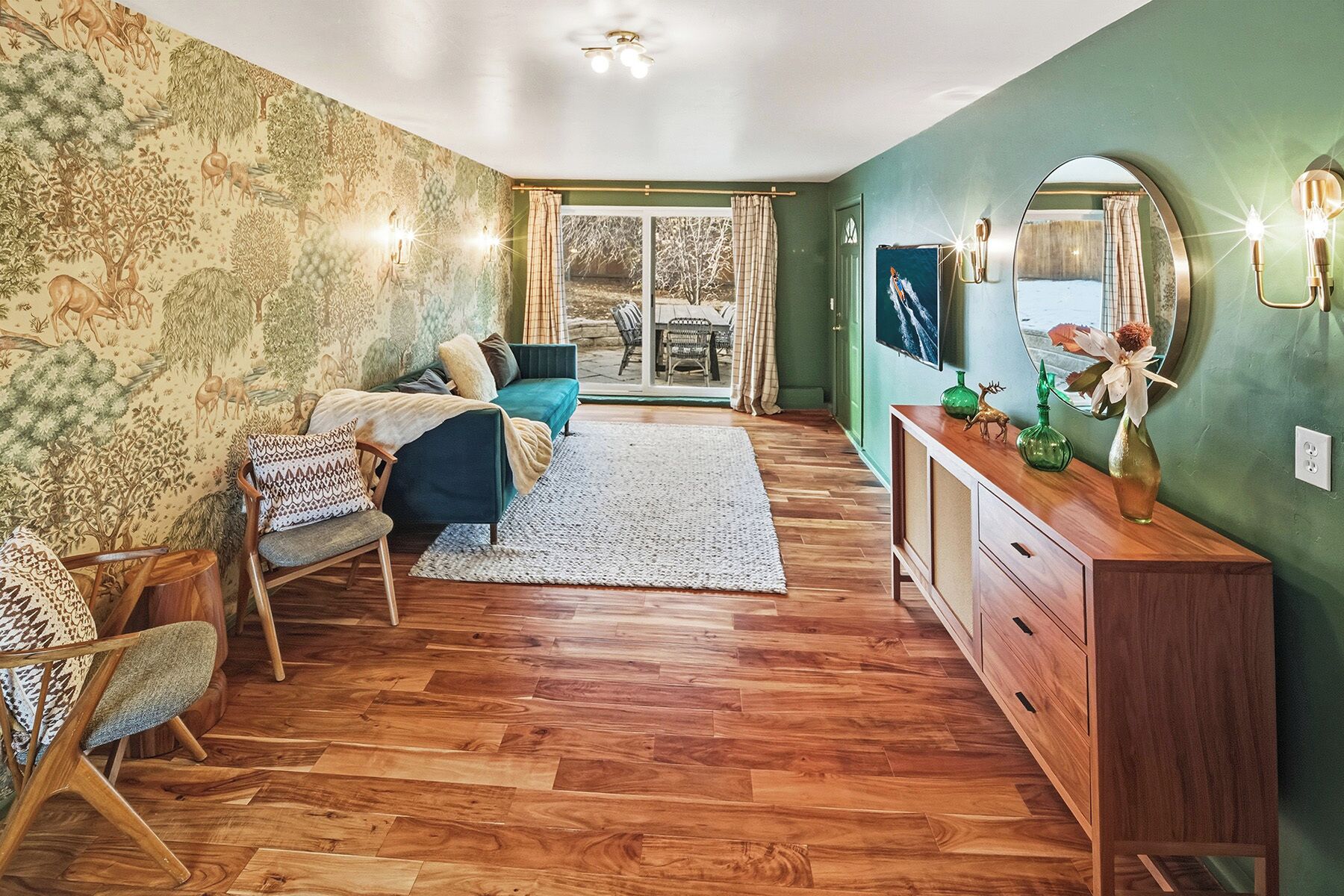 Una habitación con piso de madera, un sofá verde, un aparador de madera y un ventanal con cortinas.