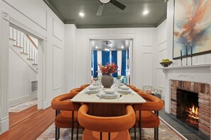 Un coin repas avec une table blanche dressée pour huit personnes, des chaises orange, une cheminée et un escalier situé à gauche.