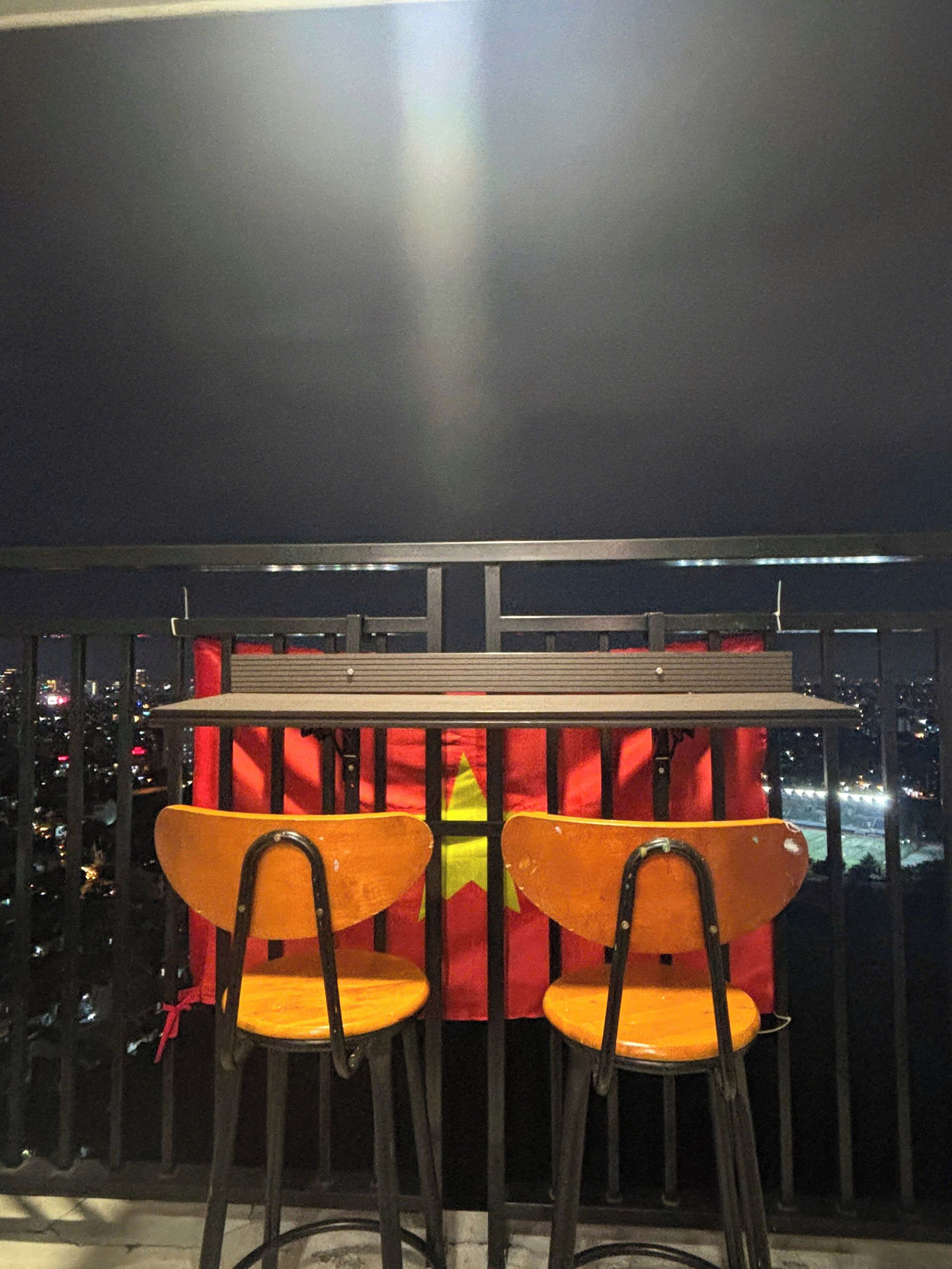A balcony with a table and two chairs, a railing, and a cityscape view at night.