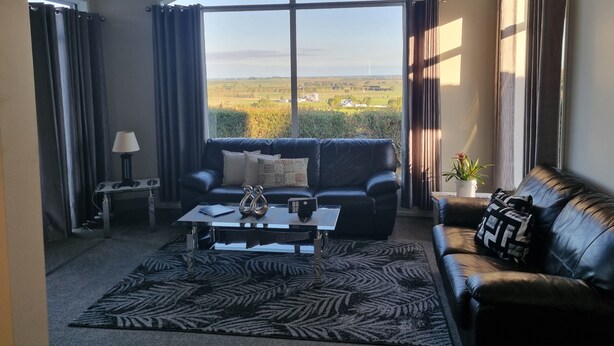 A living room with a large window, a black leather sofa, a coffee table, and a patterned rug. - Far North holiday base with panoramic coastline outlook and comfy sleepout (Kaitaia)