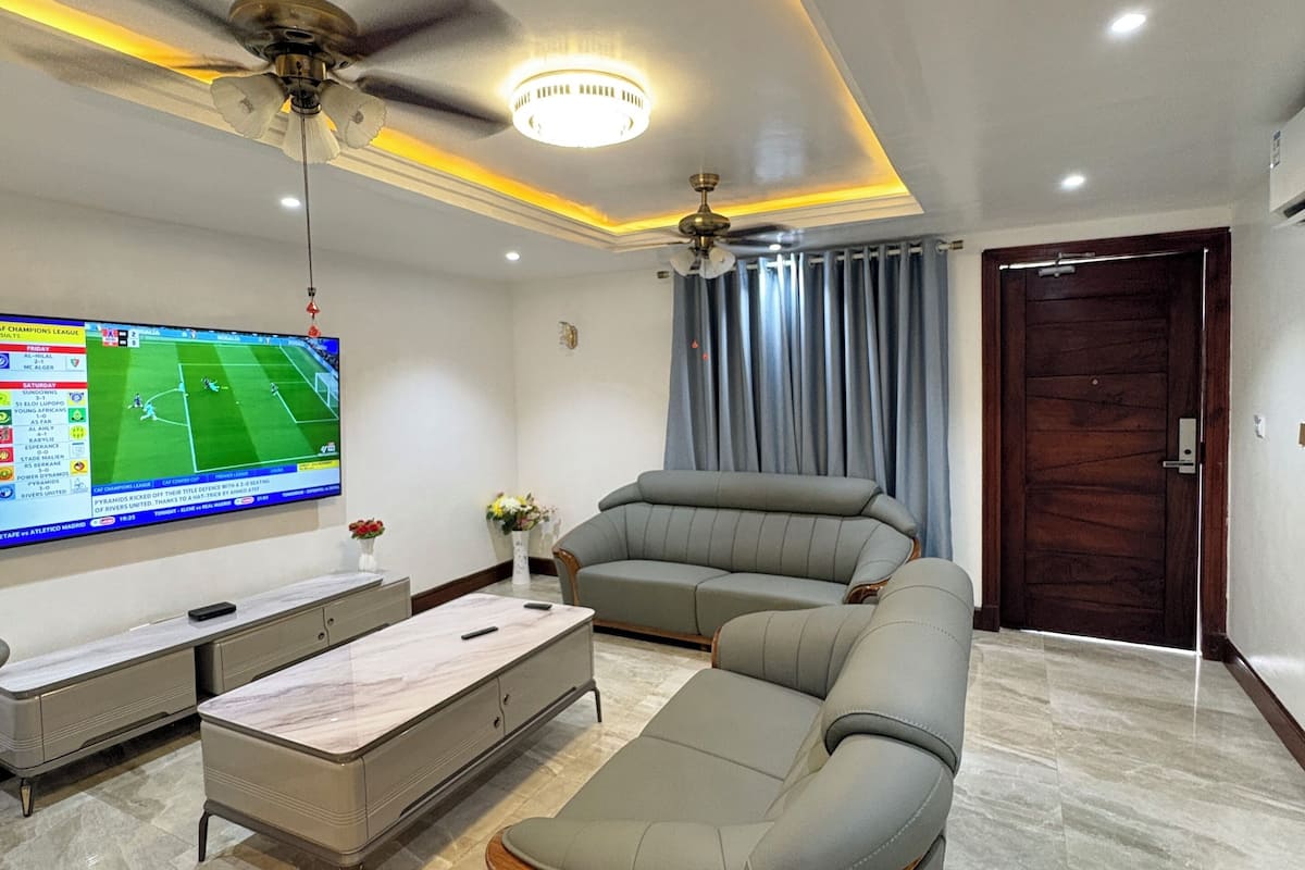 A modern living room with a large flat-screen TV, a sofa, a coffee table, and two ceiling fans.