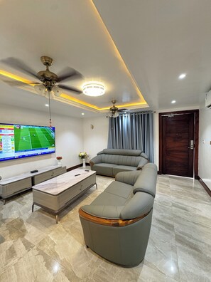 A modern living room with a large flat-screen TV, a sofa, a coffee table, and two ceiling fans. - Ridgeway Apartments (Accra)