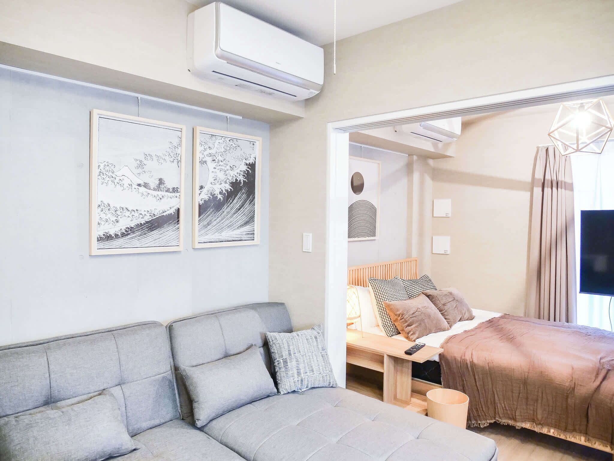 A modern living room with a grey sofa, a wooden bed with a brown blanket, and two framed artworks on the wall.