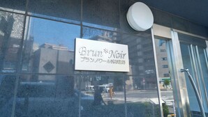 Un cartello con la scritta Brun*Noir e caratteri giapponesi, montato su un edificio con una vetrina di vetro che riflette una scena di strada.