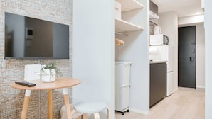 En moderne stue med et fladskærms-tv, et lille rundt bord og to stole. Der er et køkkenområde med hvide skabe og et køleskab.