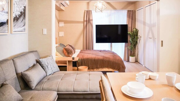 A modern hotel room with a sofa, a dining table with white cups and plates, a bed with a brown blanket, a TV, and a potted plant. - Triphome Asakusa Alpha 2 Bedrooms (Taito City)