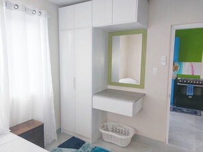 Ein Schlafzimmer mit einem Bett, einem weißen Kleiderschrank, einem Spiegel mit grünem Rahmen, einem Wäschekorb und einer kleinen Küchenzeile im Hintergrund.