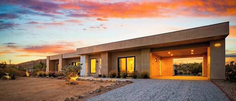 A modern two-story house with a flat roof, large windows, and a gravel driveway.