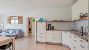 A modern kitchen with white cabinets, a wooden countertop, and a built-in oven. There is a dining area with a wooden table and chairs, and a living room with a grey sofa.