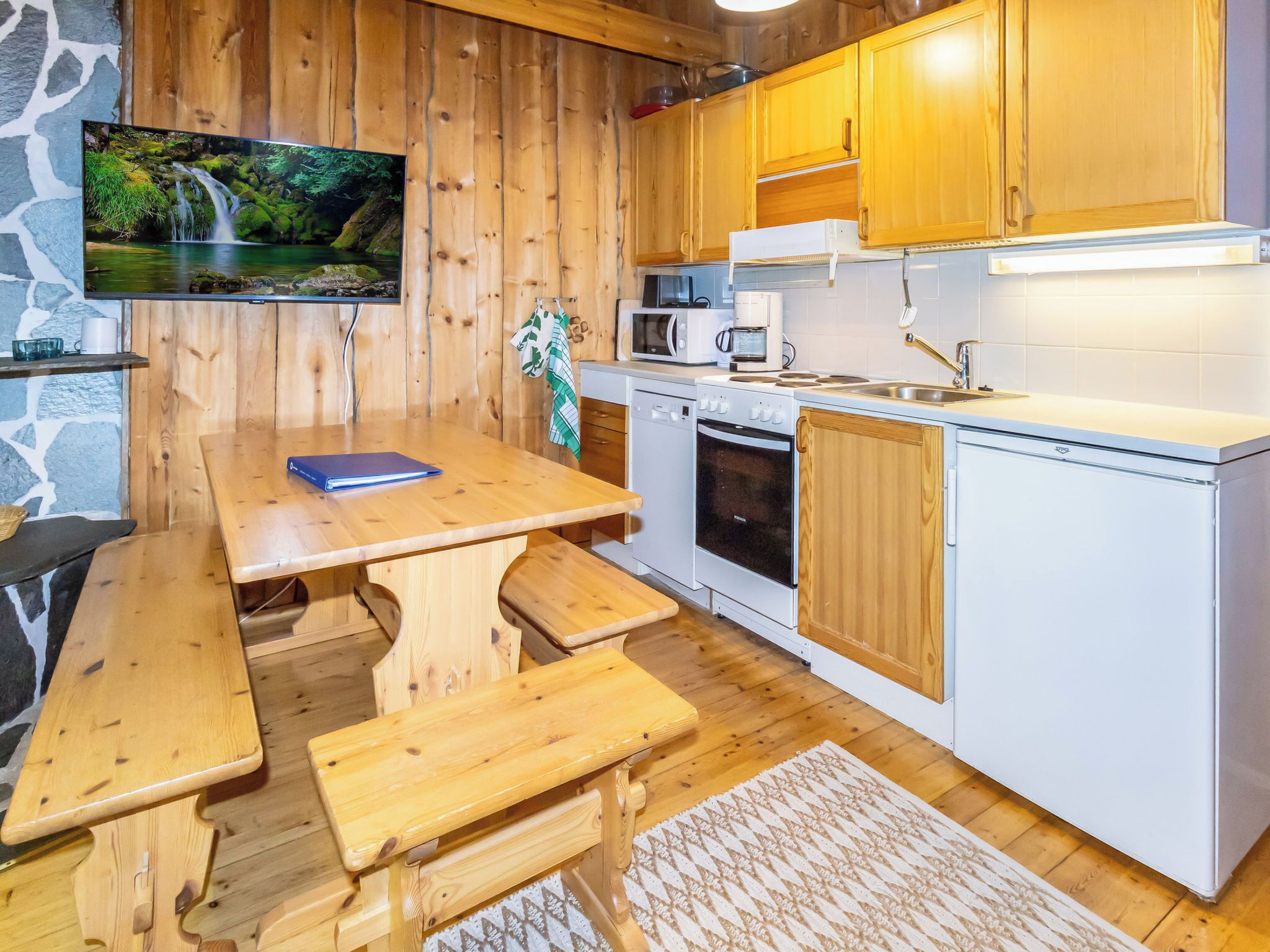 Eine gemütliche Küche mit Holdekor, einer Mikrowelle, einem Backofen und einem Spülbecken. Es gibt einen Holztisch mit Bänken und einen an der Wand montierten Fernseher.