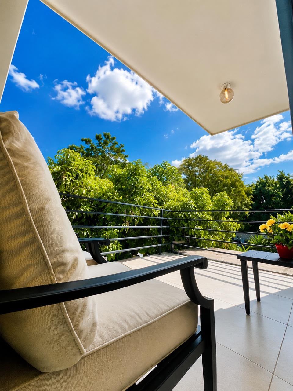 A balcony with a chair and table, a clear blue sky, and greenery in the background.