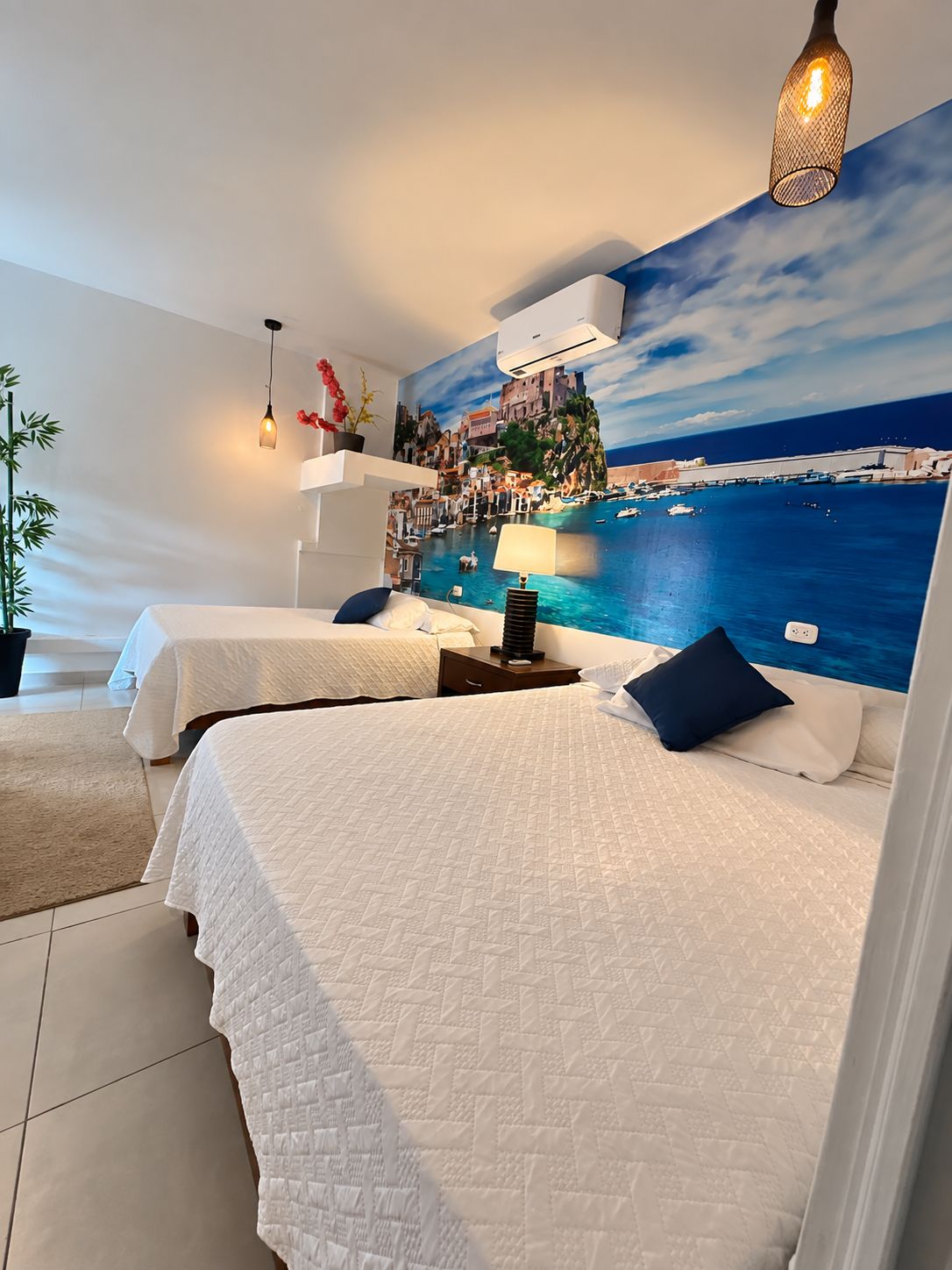 A hotel room with two beds, a mural of a coastal scene, a potted plant, and a hanging light fixture.
