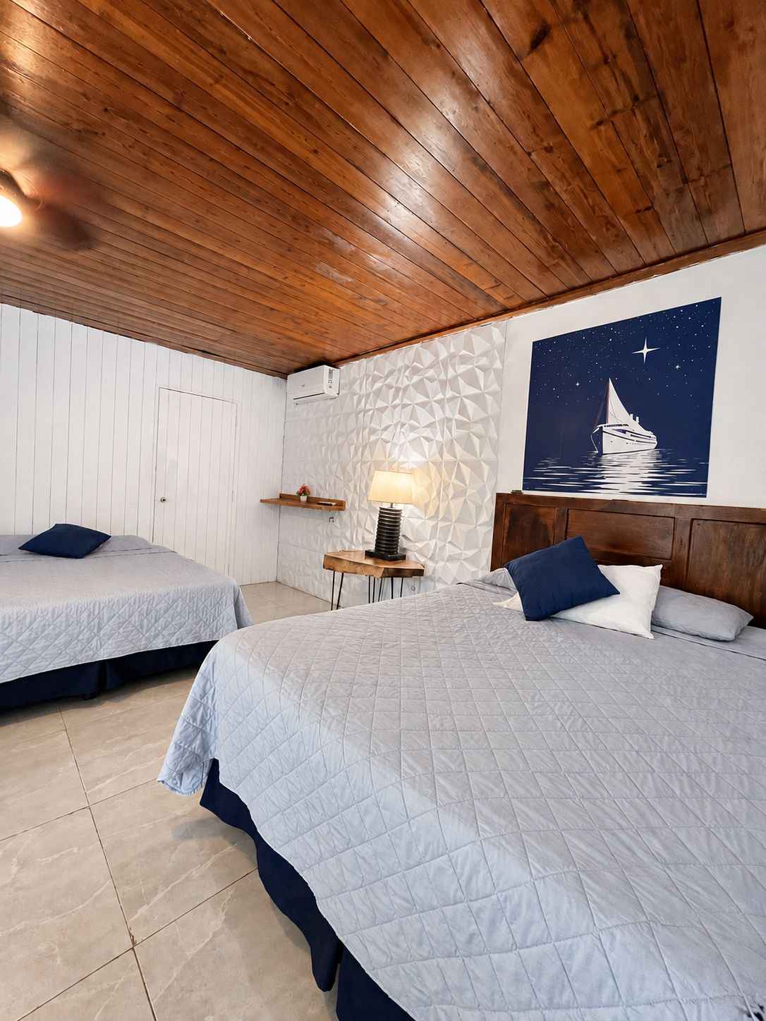Una habitación con dos camas, una mesita de noche con lámpara, aire acondicionado instalado en la pared y un cuadro con una noche estrellada.