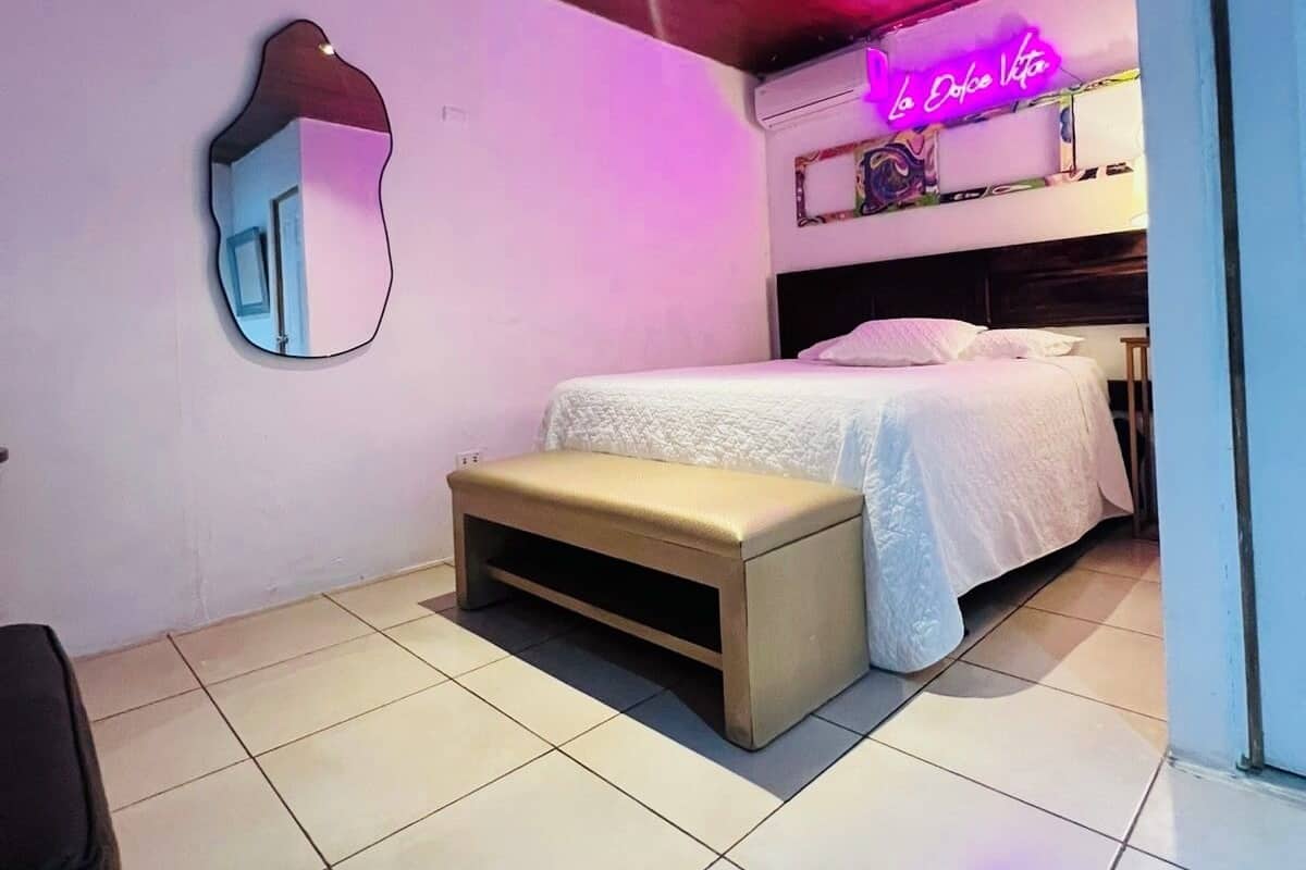 A bedroom with a bed, a bench, a neon sign, and a mirror.