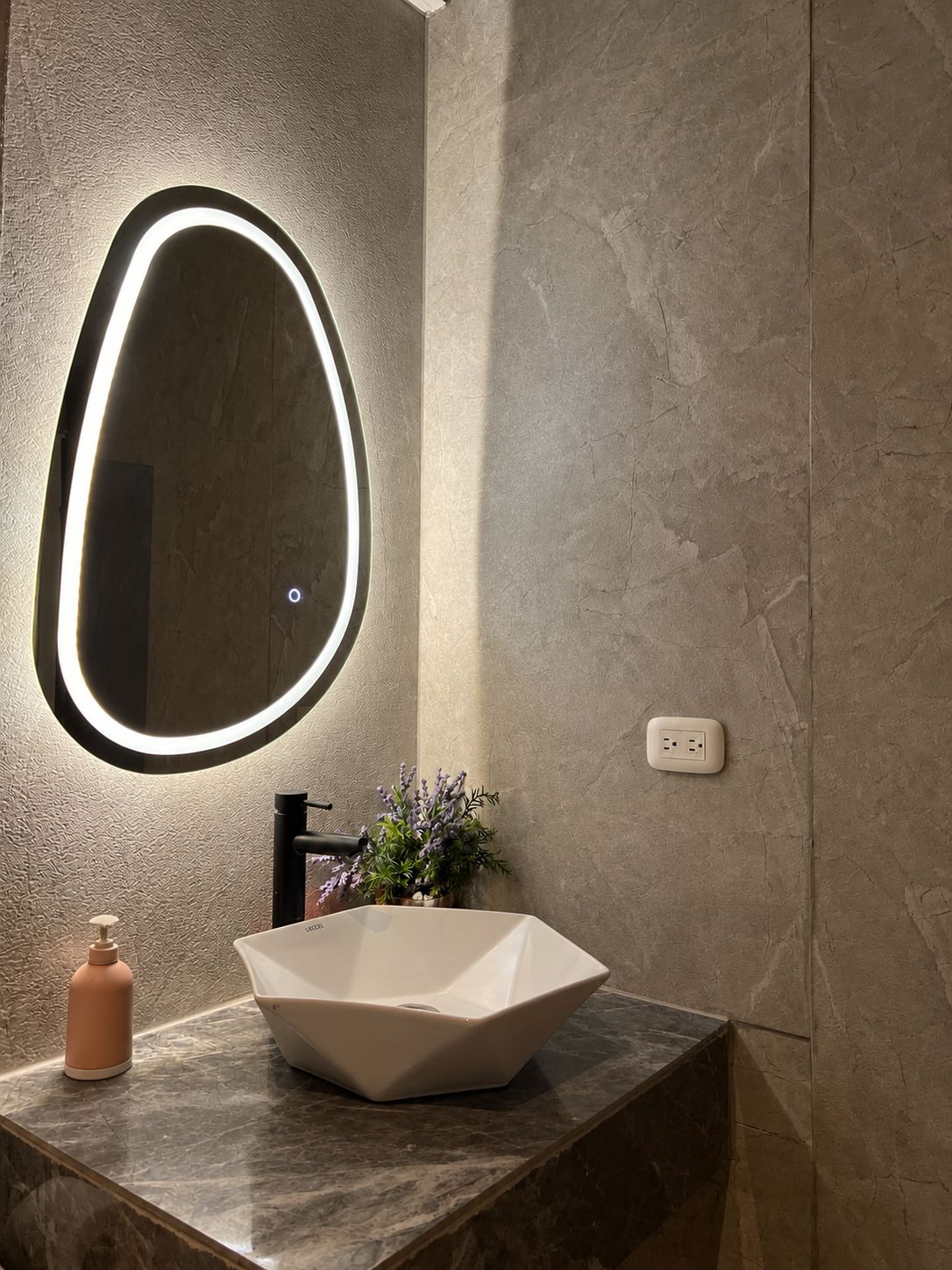 A bathroom with a modern sink, a round mirror with lighting, a potted plant, and a power outlet.