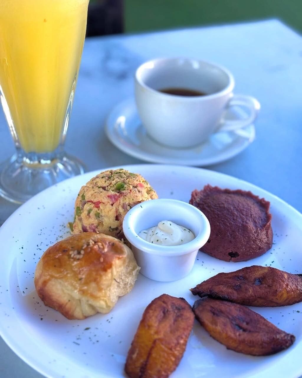 A plate with food items including a muffin, a small bowl of sauce, and roasted plantains, accompanied by a cup of coffee and a glass of orange juice.