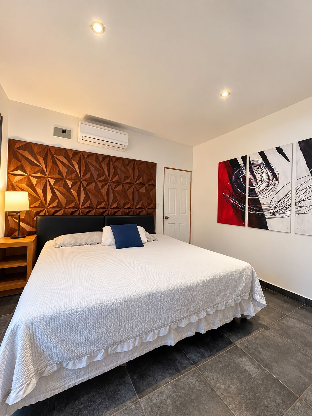 Een hotelkamer met een bed, een nachtkastje, een airconditioner, een deur en een muur met abstracte kunst.