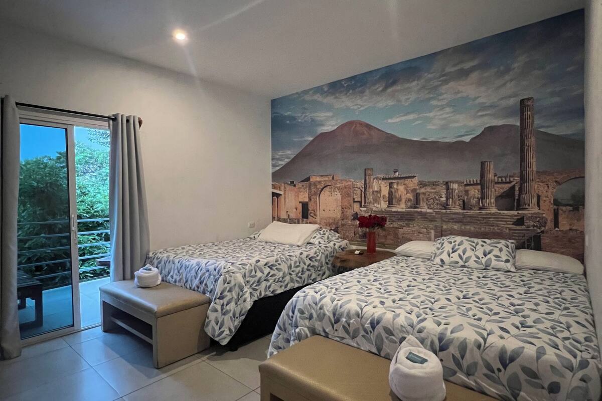 A hotel room with two beds, a large mural of a cityscape, a sliding glass door to a balcony, and a bench.