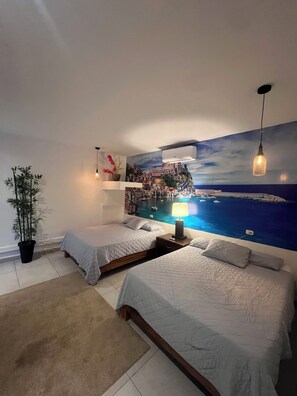 Un dormitorio con dos camas, un mural grande de paisaje costero, una planta en maceta y una lámpara colgante.