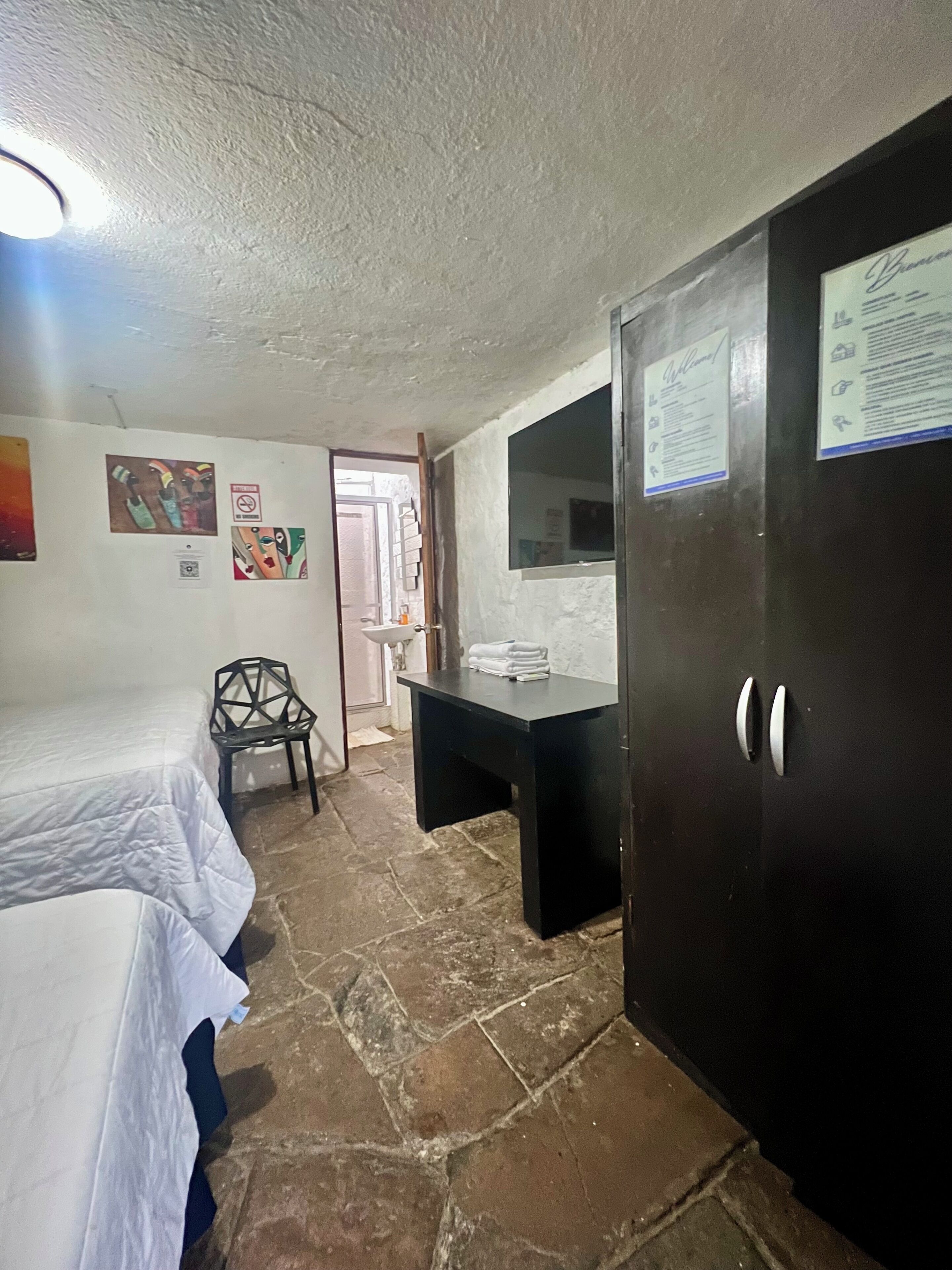Una habitación con cama, escritorio y armario. Hay carteles en las paredes y una puerta con un aviso.