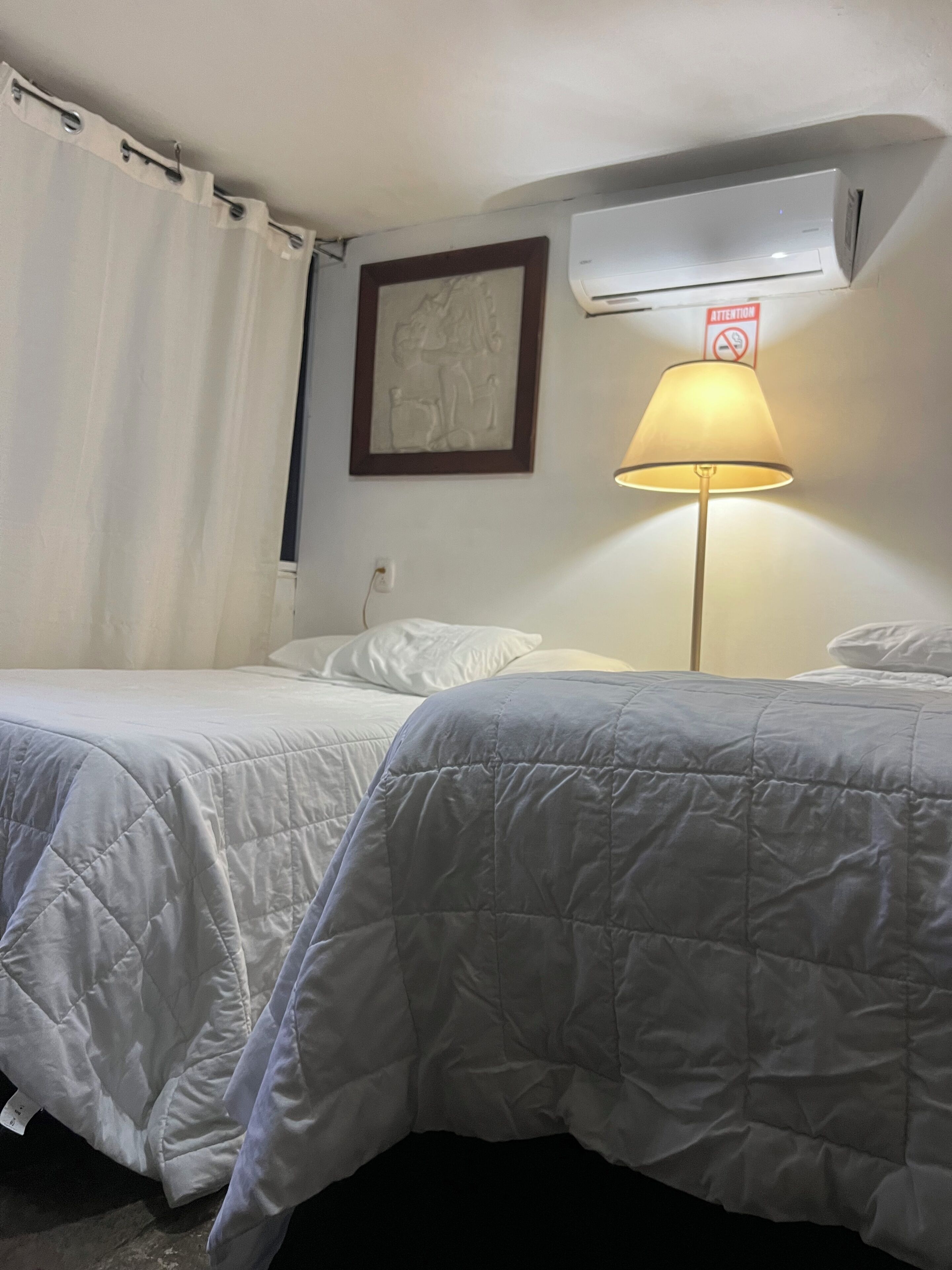 A hotel room with two beds, a lamp, an air conditioner, and a framed picture on the wall.