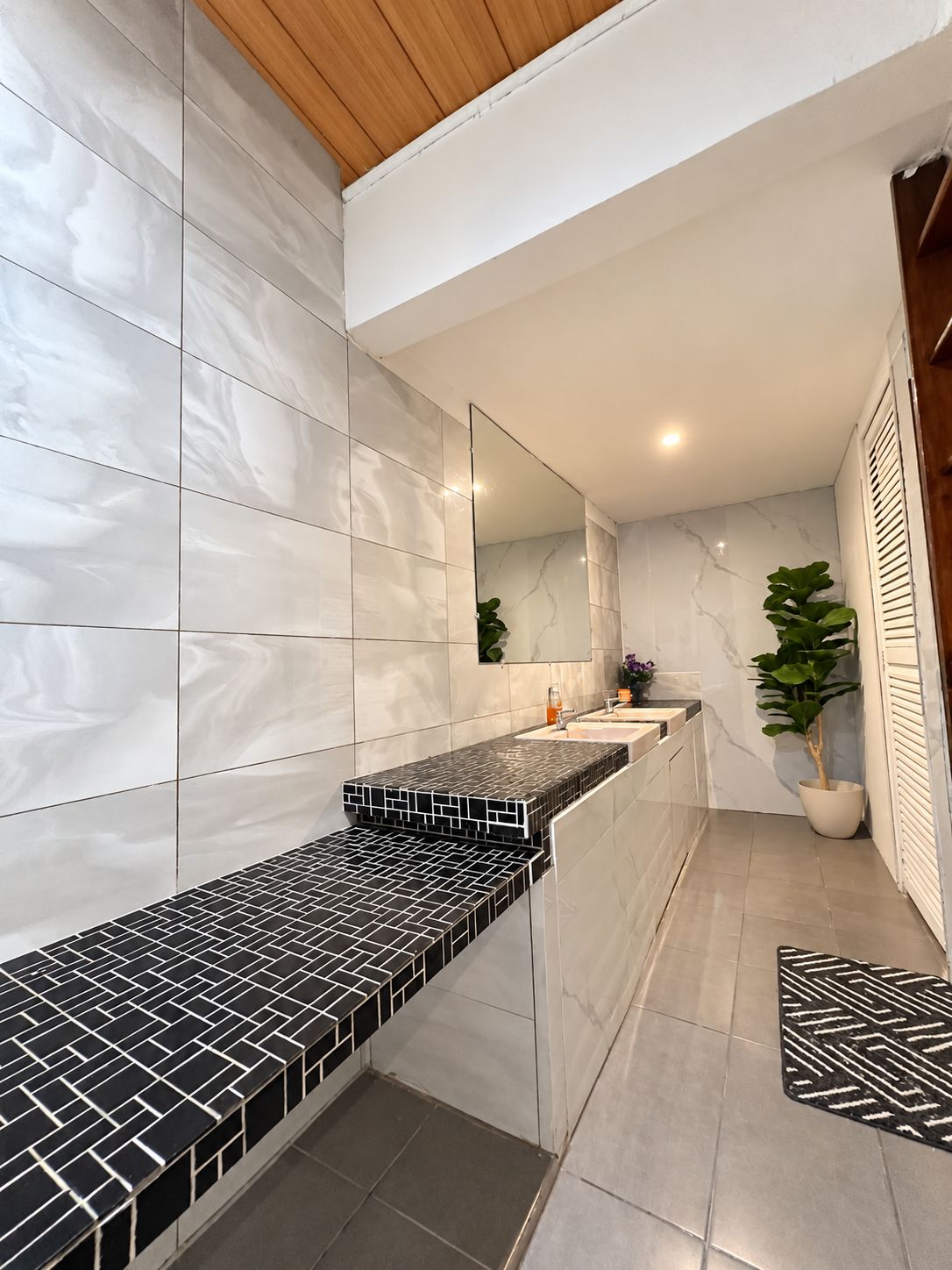 A modern bathroom with a marble wall, a black and white tiled countertop, a mirror, and a potted plant.