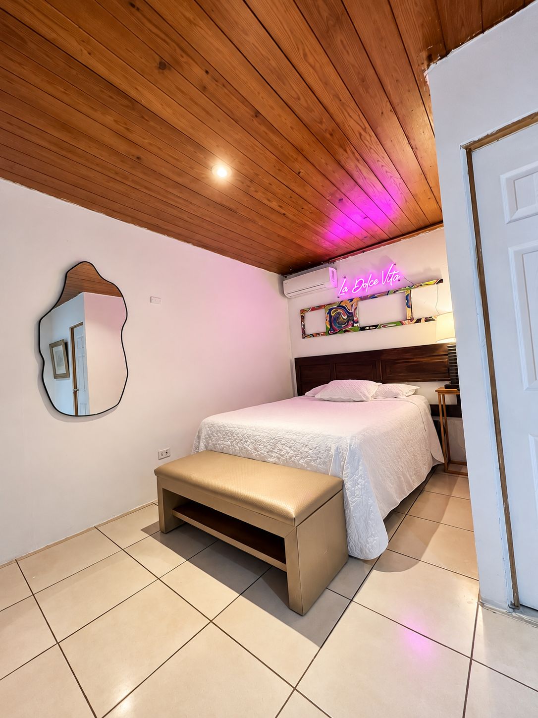 A bedroom with a wooden ceiling, a bed with white bedding, a bench, a neon sign, and a mirror.