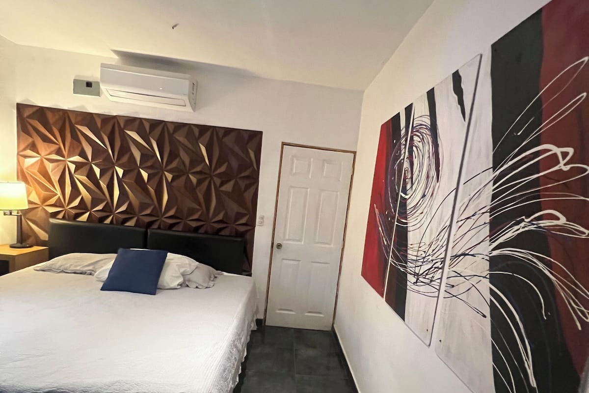 A hotel room with a bed, a wall-mounted air conditioner, a door, and abstract wall art.