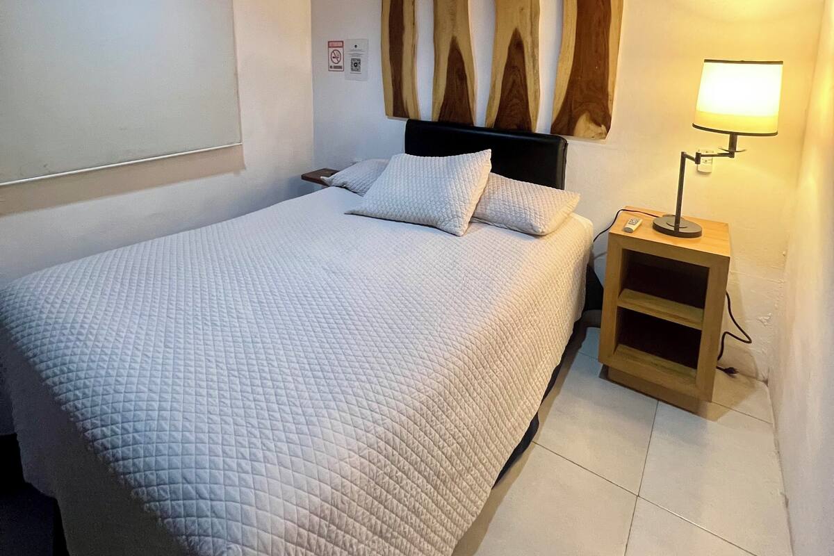 A bedroom with a bed, a nightstand with a lamp, wooden wall decor, and a white ceiling.