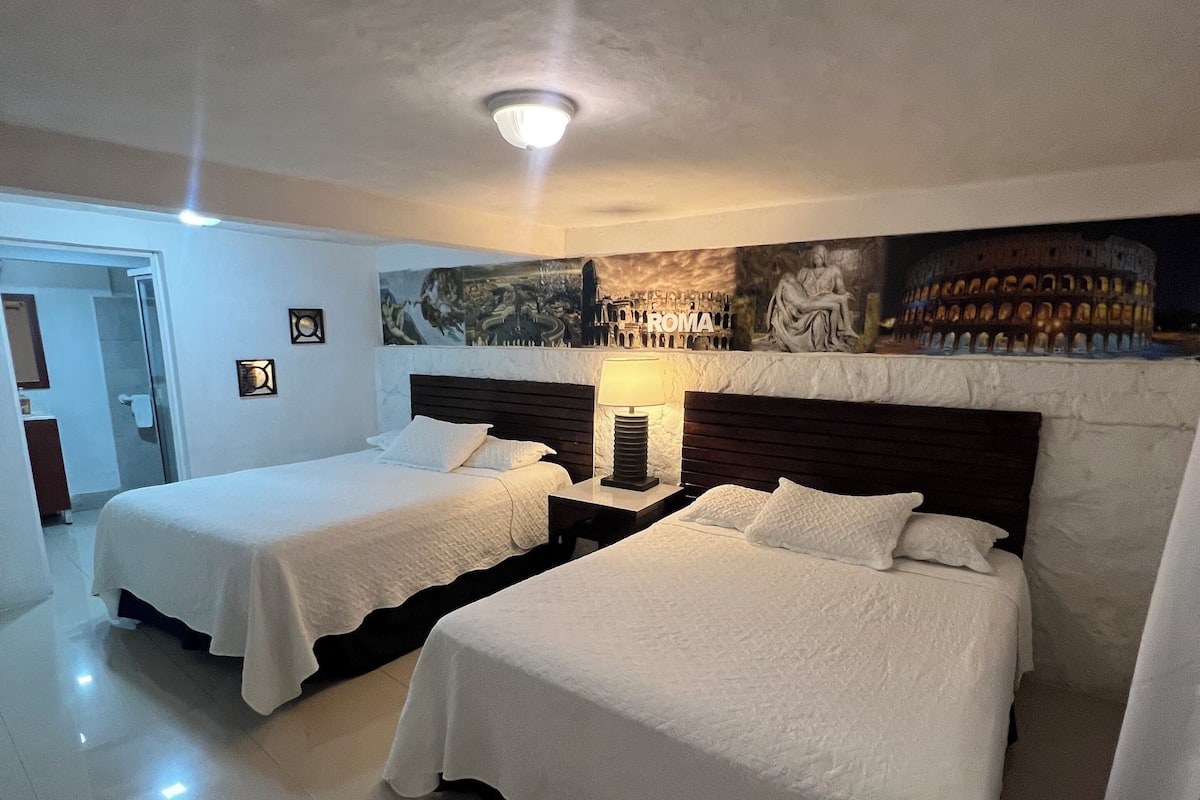 A hotel room with two beds, a large mural of the Colosseum, and a modern bathroom visible through an open door.