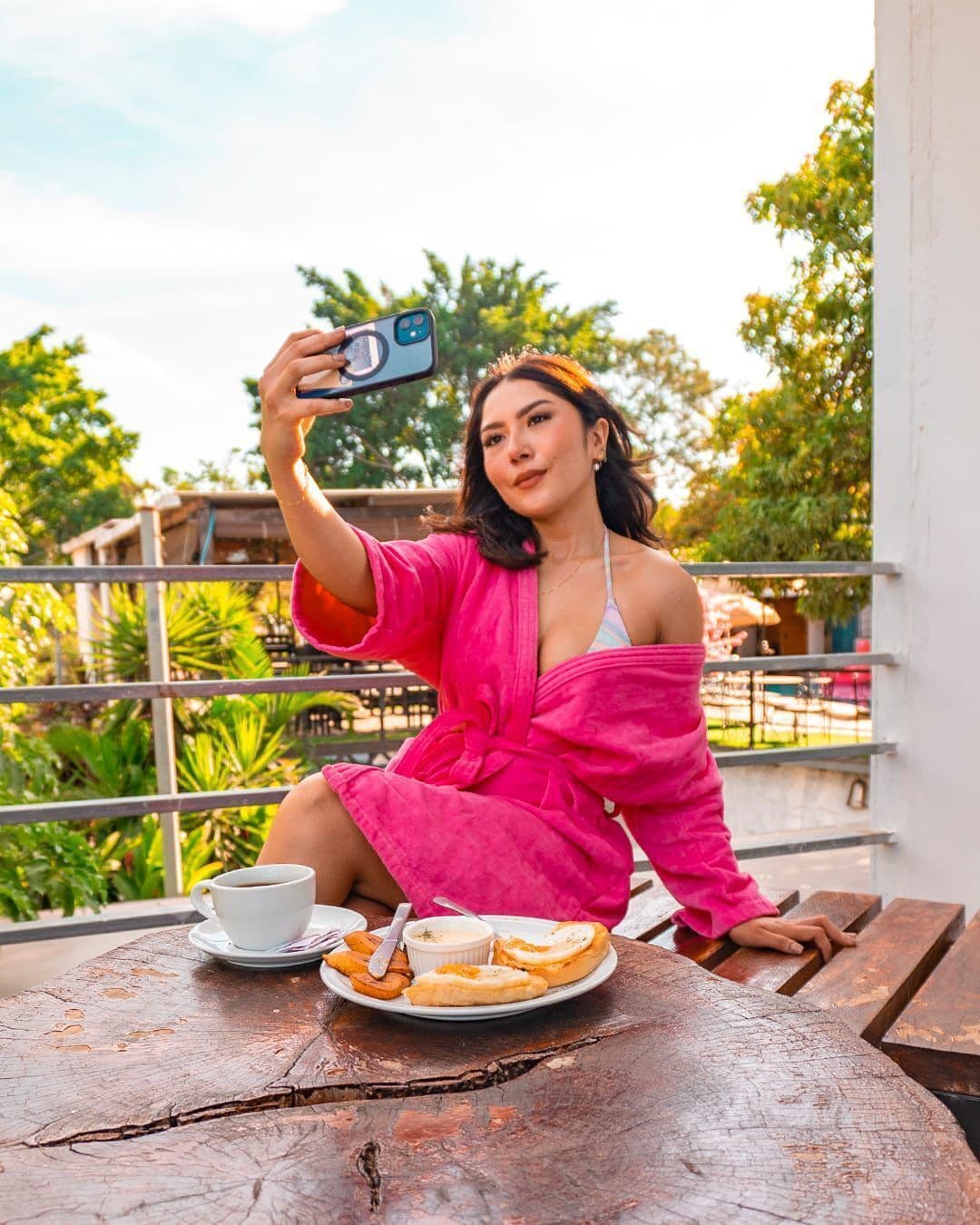 Uma pessoa tira uma selfie em uma mesa ao ar livre, com um prato de comida e uma xícara de café.