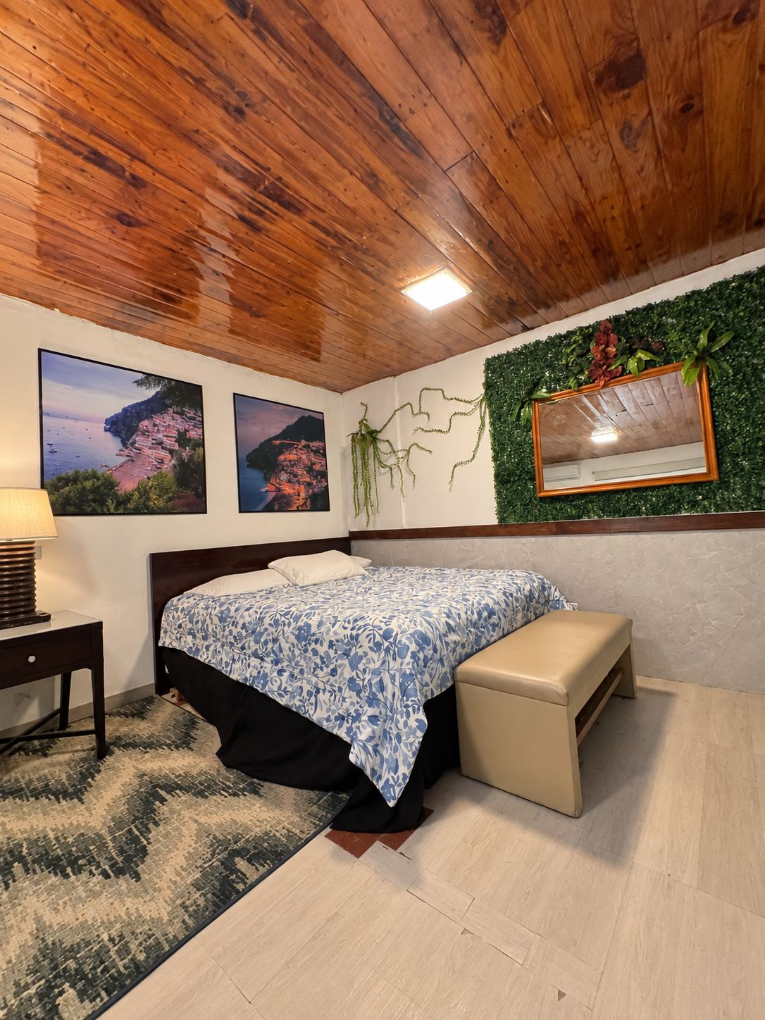Ein Schlafzimmer mit Holzboden, einem Bett mit gemustertem Überwurf, einer Bank, einem Nachttisch, einer Lampe und zwei gerahmten Bildern an der Wand.