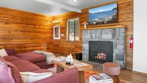 L’intérieur chaleureux d’un chalet avec une cheminée en pierre, des murs en bois, un grand téléviseur fixé au-dessus de la cheminée et un coin salon confortable avec un canapé et une table.