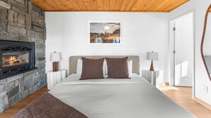 Une chambre avec une cheminée en pierre, un lit avec deux oreillers, une table de chevet avec une lampe et un tableau encadré au mur.