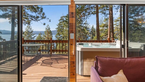 Une terrasse en bois avec un jacuzzi, une table et des chaises, et une vue sur les arbres et les montagnes.