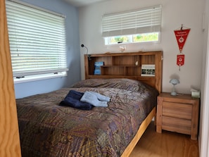 Un dormitorio con cama, mesita de noche, lámpara, estantería con libros y ventana con persianas.