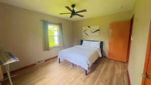 Una camera da letto con un letto, una ventola a soffitto, una finestra con tende, una porta e un'opera d'arte appesa al muro.