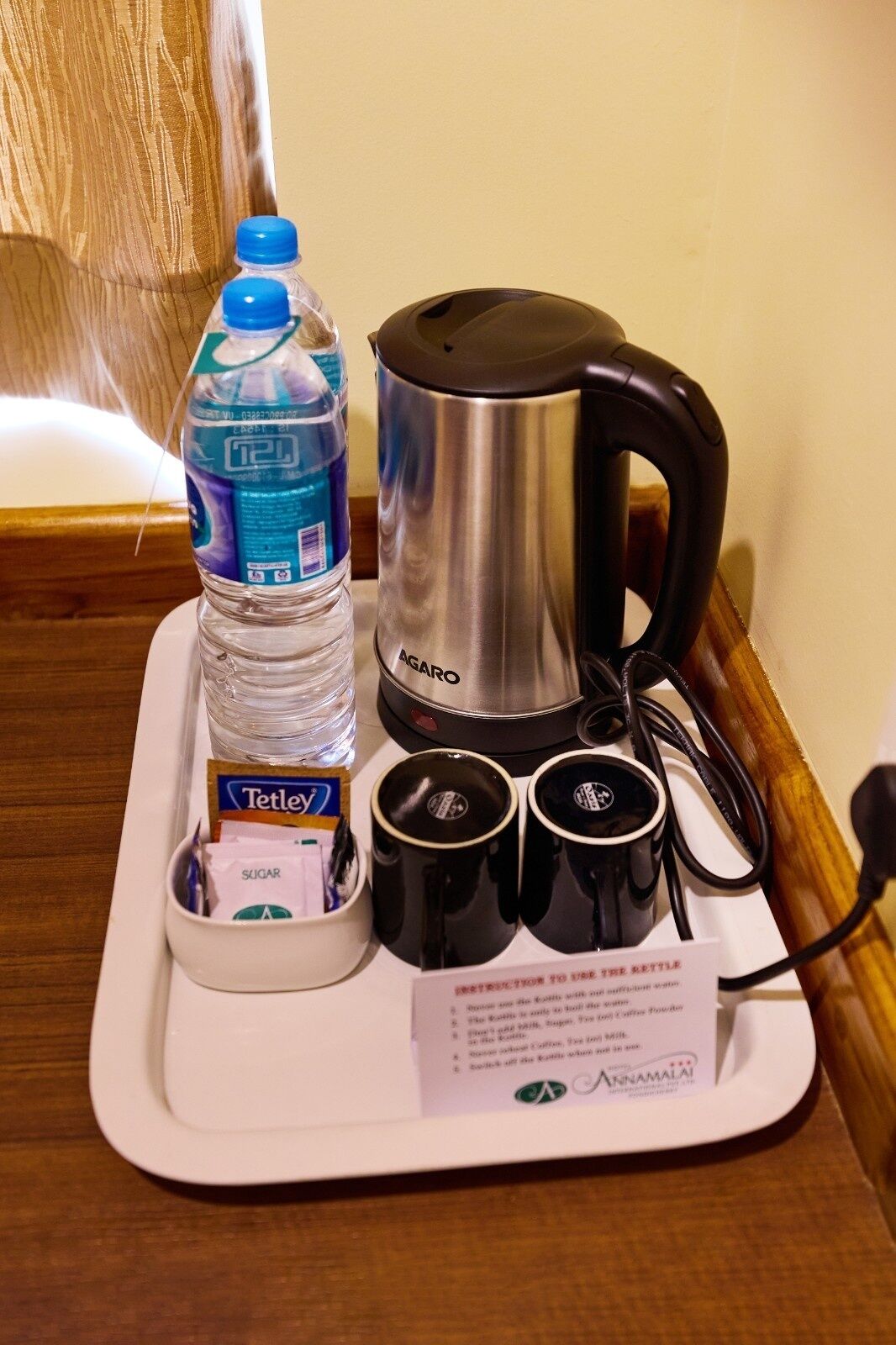 A tray with a water bottle, a kettle, sugar packets, and two black mugs on a hotel table.