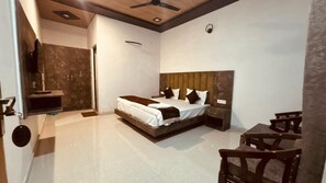 A bedroom with a bed, a wooden headboard, a bedside table, a TV mounted on the wall, and a ceiling fan. - Rajaji Retreat Resort (Ganga Bhogpur Talla)