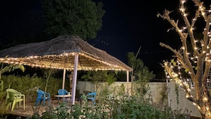 Un espace extérieur aménagé avec des sièges et un toit de chaume, illuminé de guirlandes lumineuses et entouré de verdure.