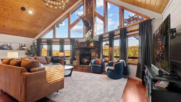 A spacious living room with a stone fireplace, large windows, and a television. - The Elk's Club - Views, Firepit, Hot Tub, Pickle Ball & Game Rm (Blue Ridge)