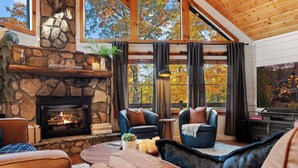 A cozy living room with a stone fireplace, leather sofas, and large windows offering a view of autumn trees. - The Elk's Club - Views, Firepit, Hot Tub, Pickle Ball & Game Rm (Blue Ridge)