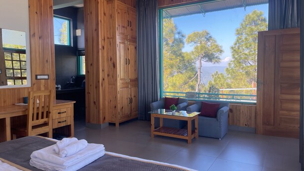 A wooden room with a bed, a desk, a sofa, and a large window offering a view of trees. - Kasar Escape (Almora)