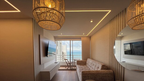 Een moderne woonkamer met een bank, televisie en eethoek, met uitzicht op een balkon en de oceaan.
