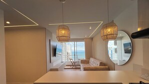 Een moderne hotelkamer met een groot raam dat uitzicht biedt op de oceaan en gebouwen. Er is een bank, een aan de muur bevestigde televisie en een ronde spiegel.